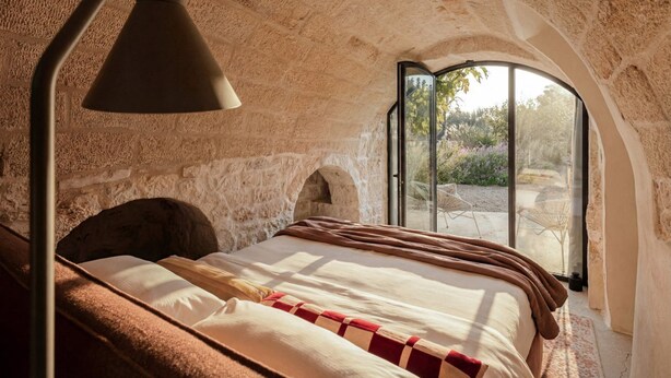 Un dormitorio con pared de piedra, una cama con manta de cuadros rojos y blancos y un ventanal con vistas a la vegetación.