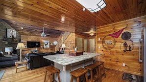Interior de una cabaña con techo de madera, chimenea de piedra, televisor grande y zona de cocina con encimera de mármol y taburetes.