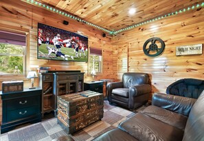 A cozy room with a wooden ceiling, a large TV screen, a leather sofa, a small table, and a wall clock.