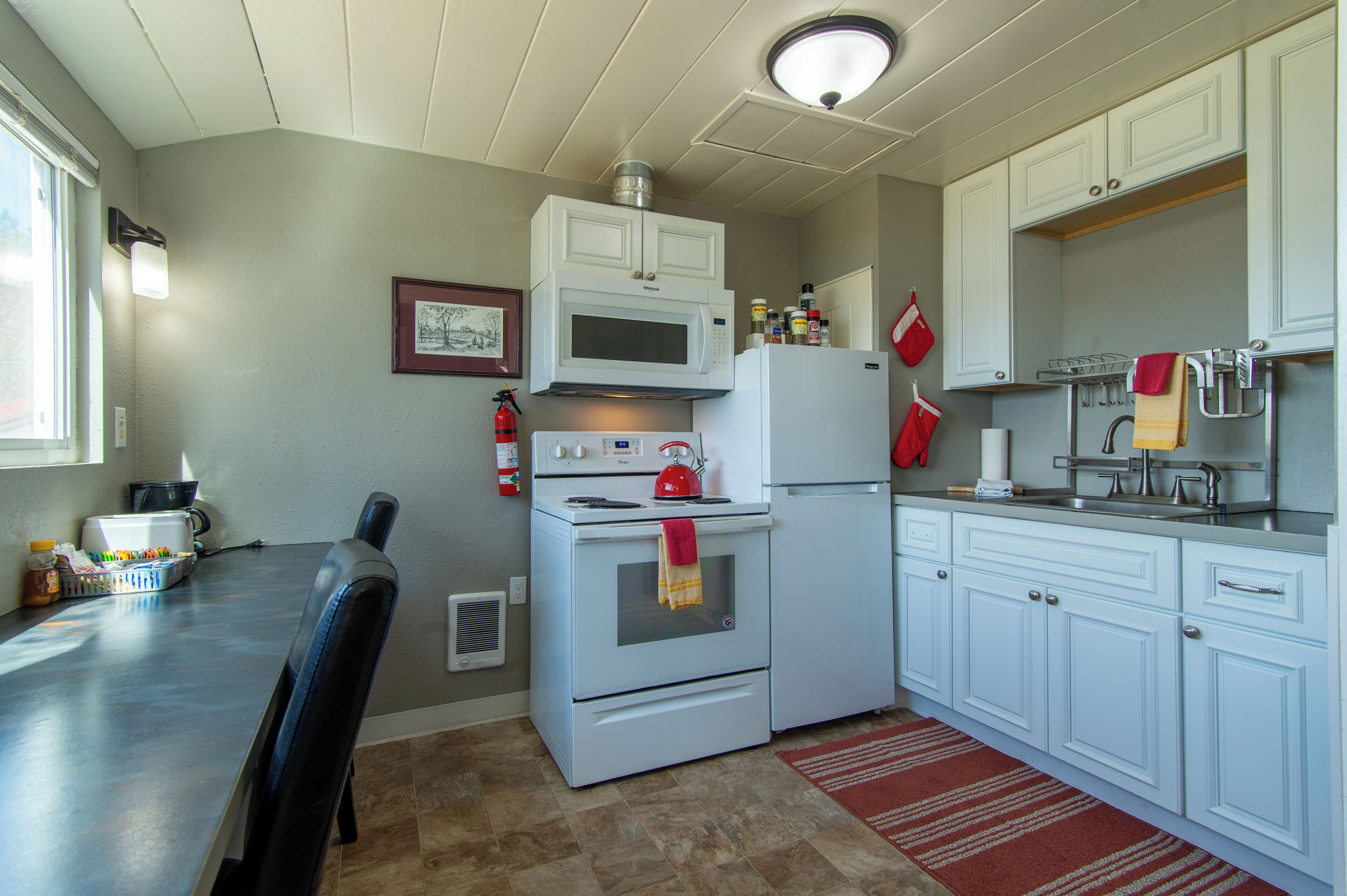 A compact kitchen with white cabinets, a microwave, a stove, and a refrigerator. There is a dining table with chairs and a red rug on the floor.