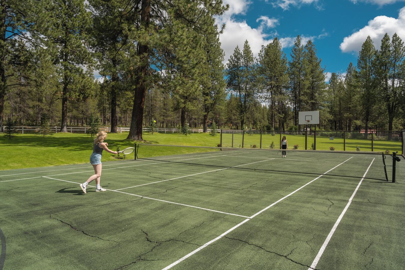 Un court de tennis où s’affrontent deux joueurs, entouré d’arbres et agrémenté d’un panier de basket.