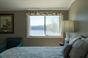 Un dormitorio con cama, una silla, una ventana con vista a un lago y una lámpara en una mesita de noche.
