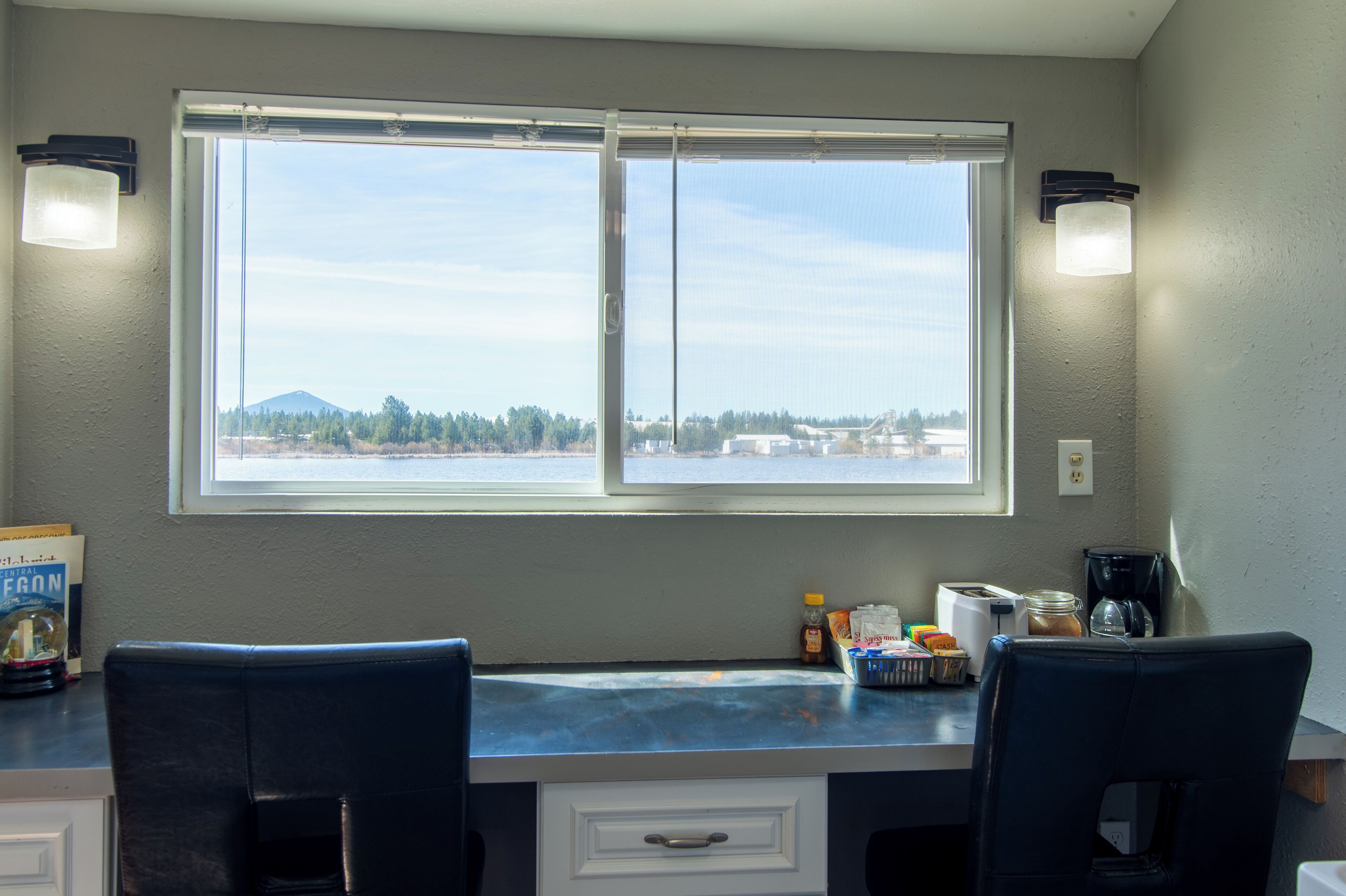 A kitchen with a large window, two chairs, a countertop with various items, and a view of a landscape with trees and a mountain.