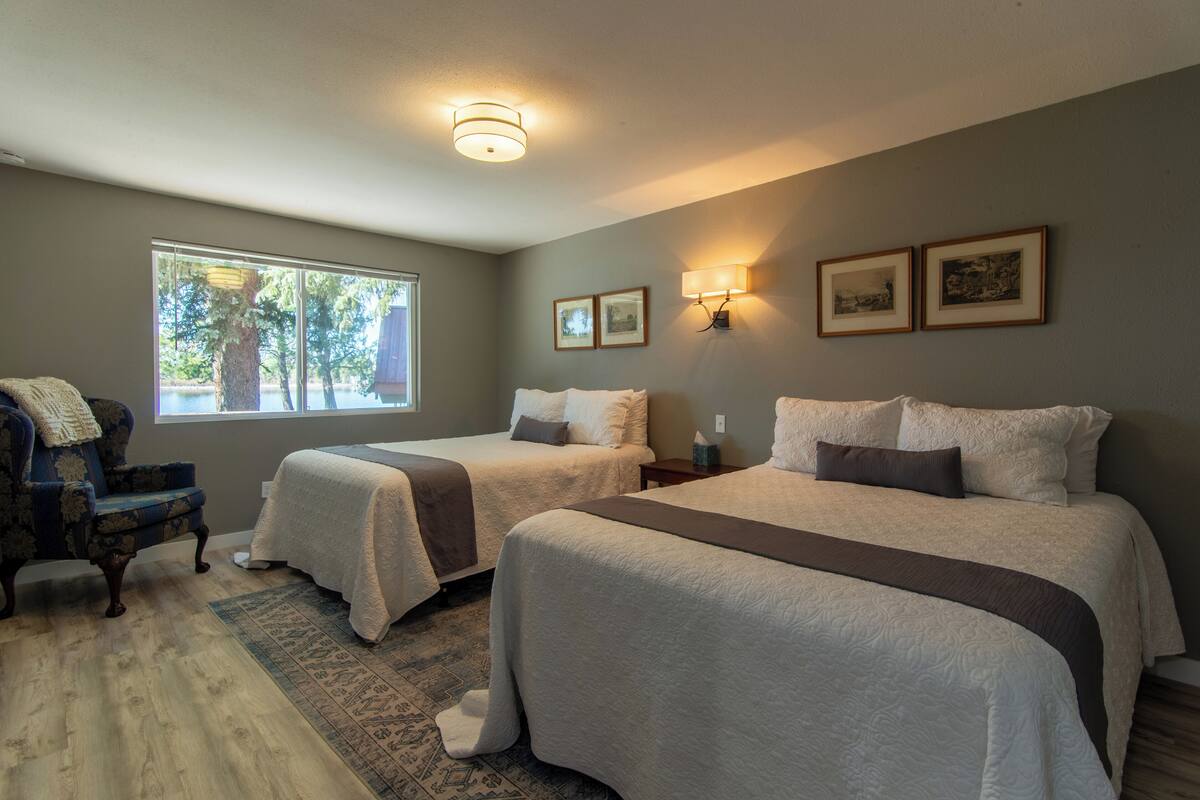 A bedroom with two beds, a chair, a window with a view of trees, and framed pictures on the wall.