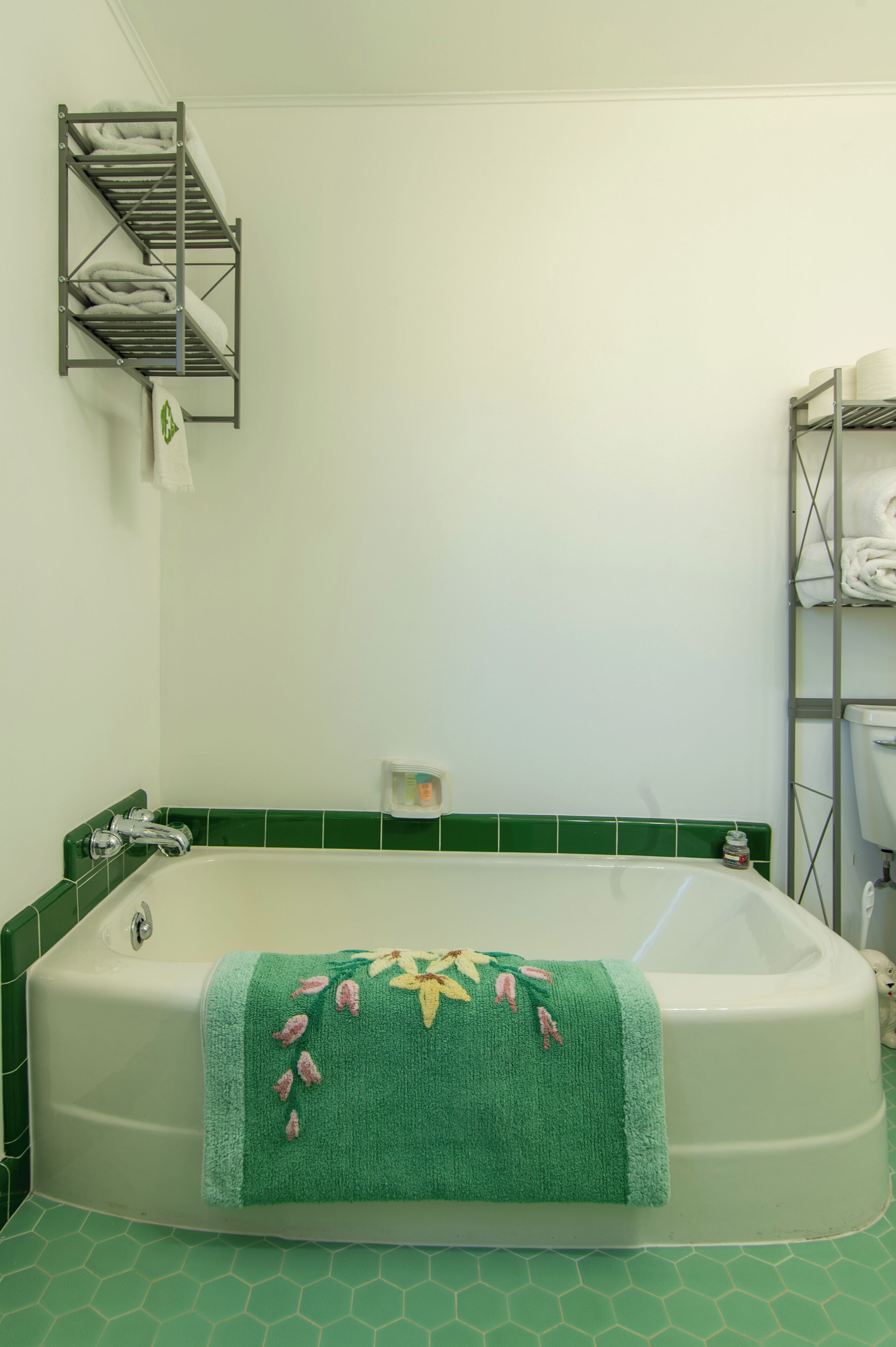 A bathroom with a green-tiled floor, a white bathtub with a green towel, and a metal shelf with towels.