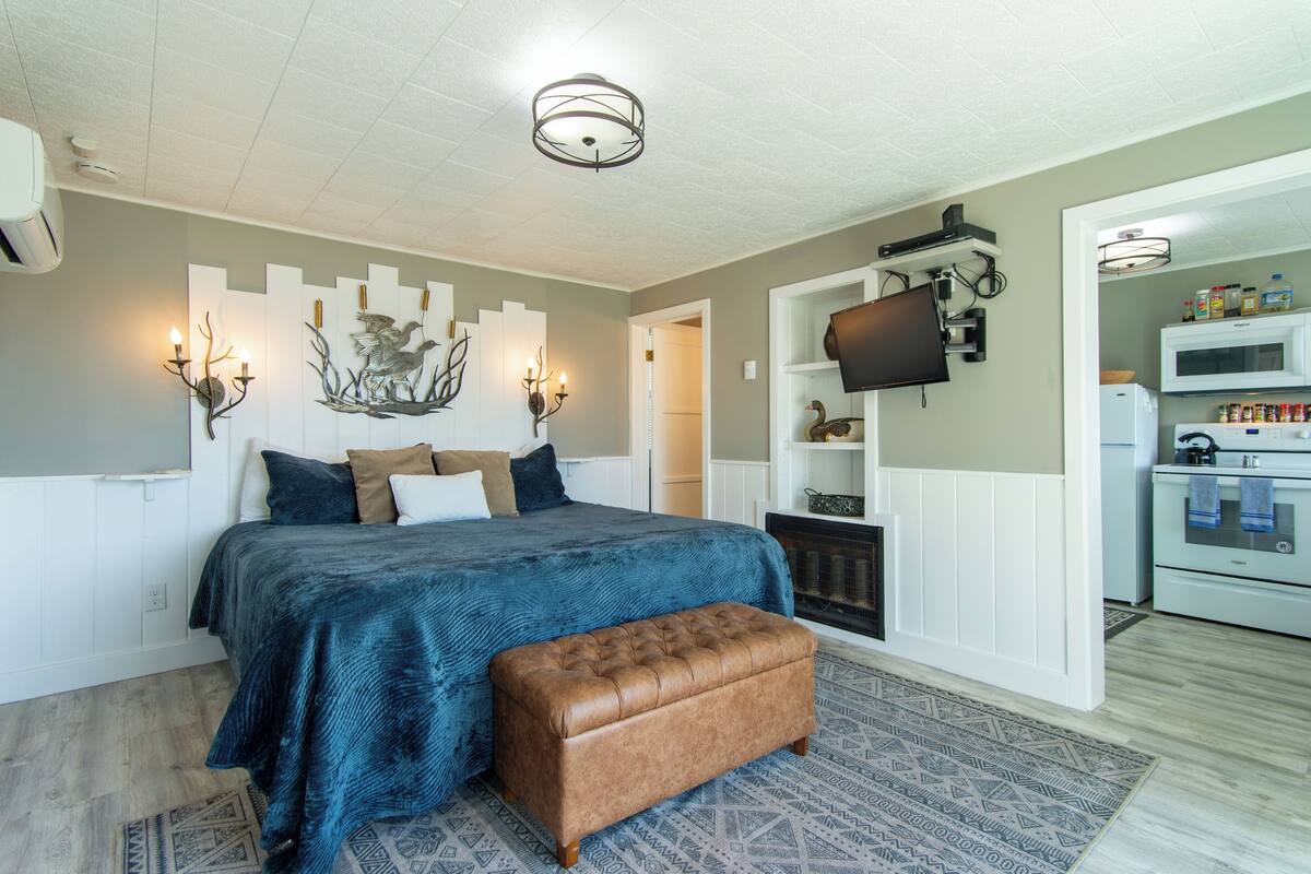 A bedroom with a bed, a bench, a fireplace, a television, and a kitchenette.
