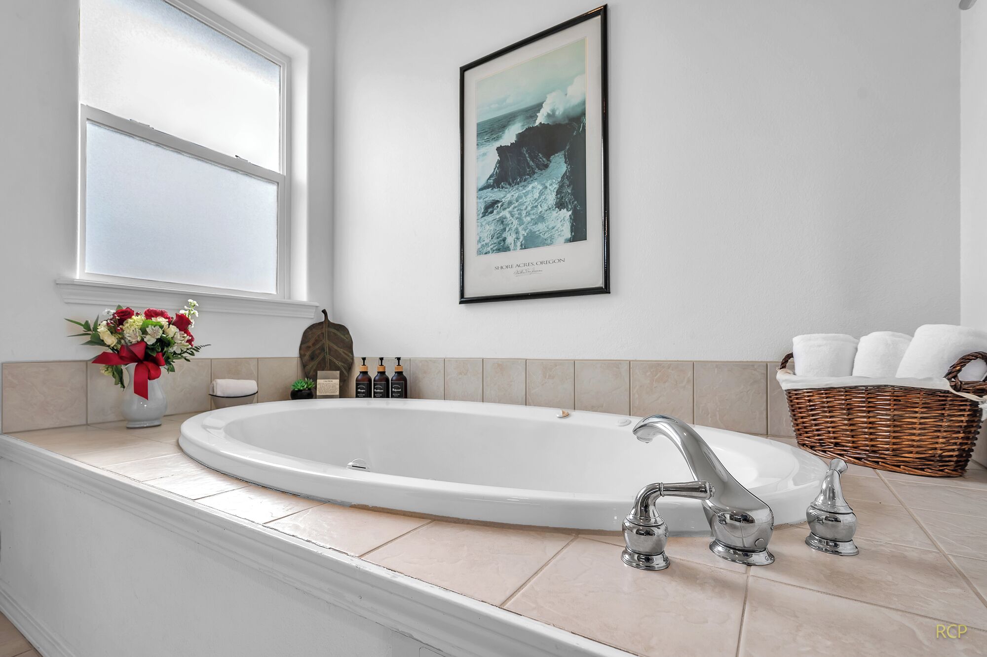 A modern bathroom with a large bathtub, a framed photograph on the wall, a vase of flowers, and a wicker basket with towels.