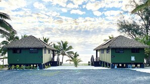 Two thatched-roof buildings labeled "Light 1" and "Light 2" on a gravel beach with palm trees and a clear sky. - Manta-Ray Beach Unit 2 (Rarotonga)