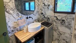 A bathroom with a marble wall, a round mirror, a sink, a window, and a trash bin. - Manta-Ray Beach Unit 2 (Rarotonga)