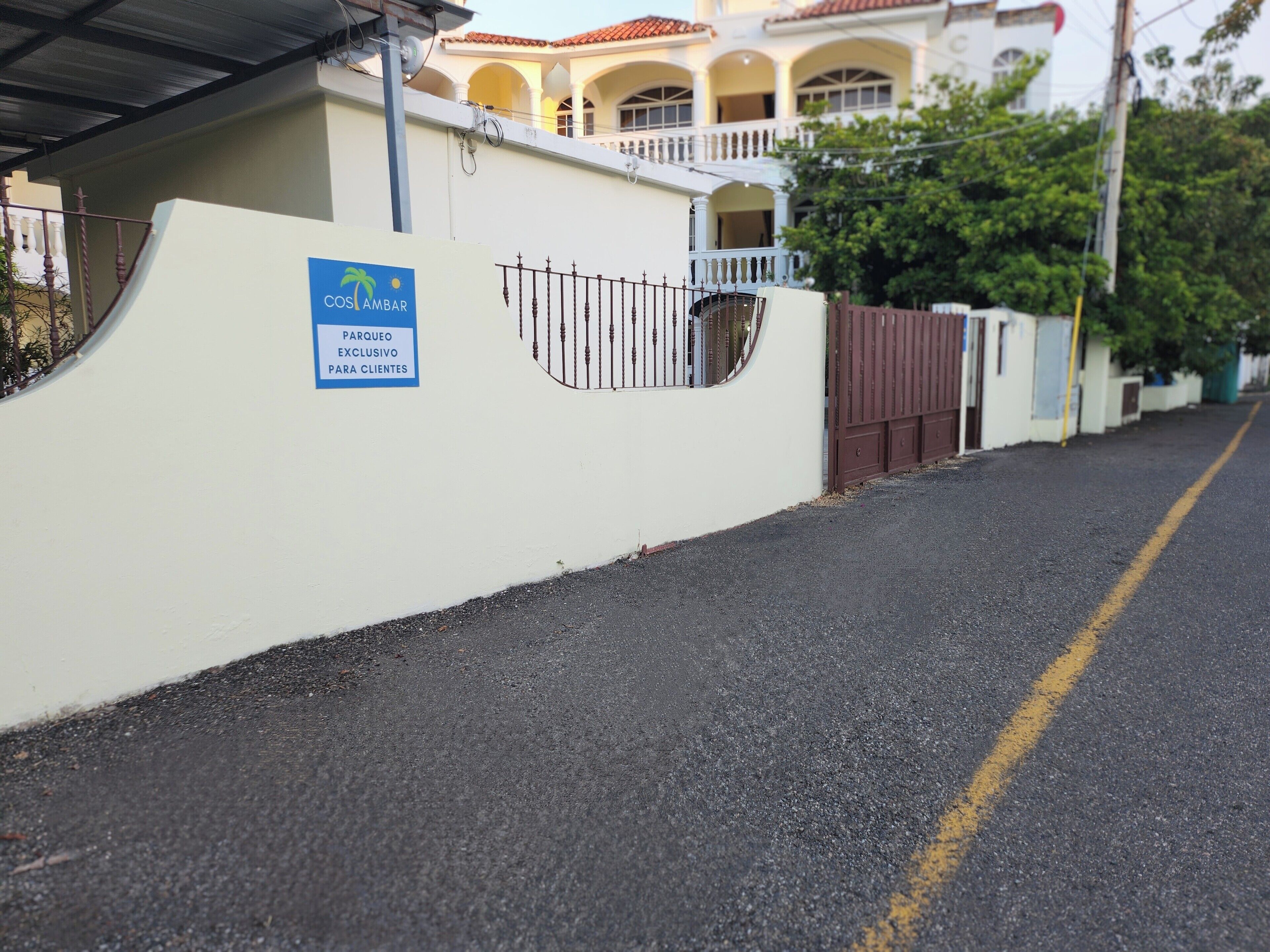 Une entrée fermée par un portail, sur laquelle on peut lire l’inscription « COSI LAMBAR, PARC DE STATIONNEMENT RÉSERVÉ AUX CLIENTS », et, au fond, un bâtiment à la façade blanche et au toit recouvert de tuiles rouges.