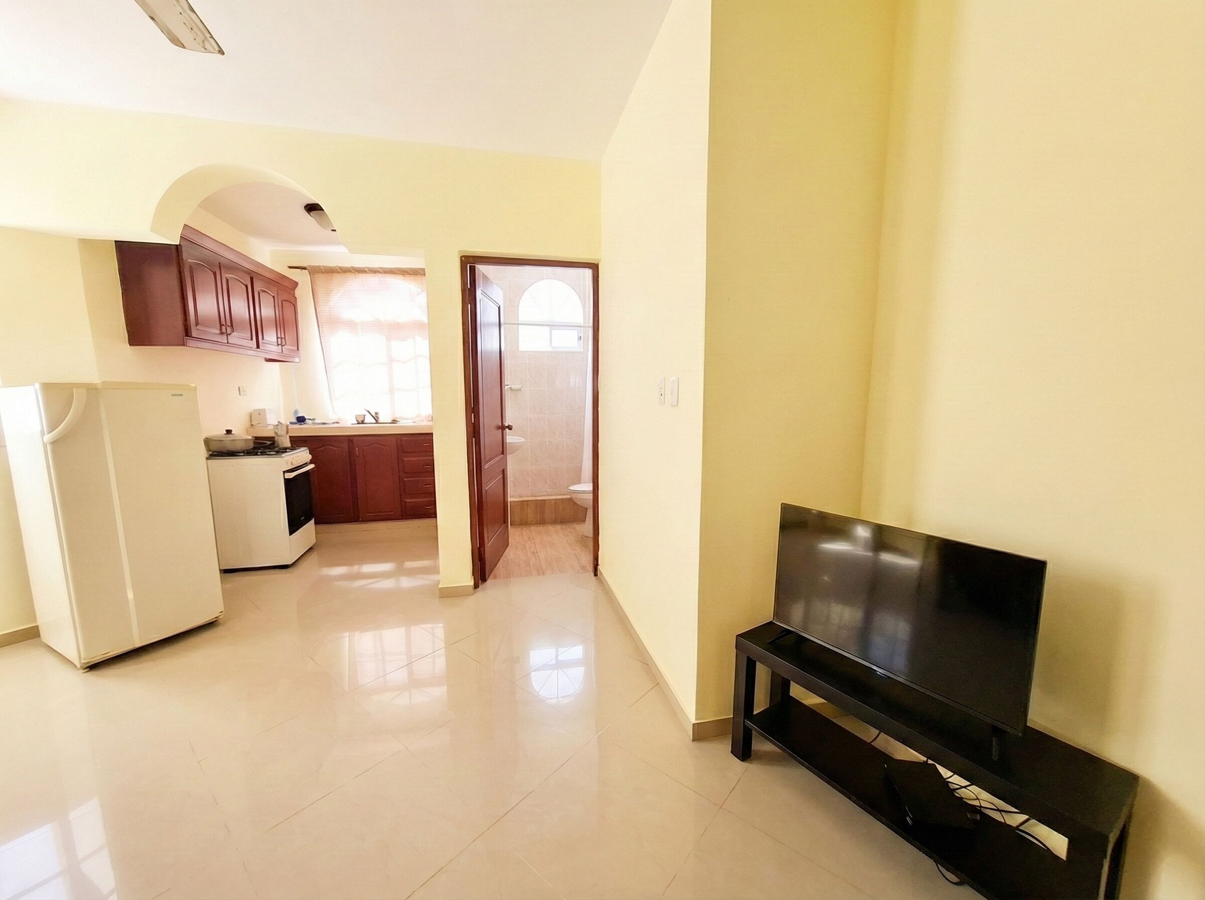 A modern living room with a flat-screen TV on a stand, a kitchen area with cabinets and appliances, and a doorway leading to another room.