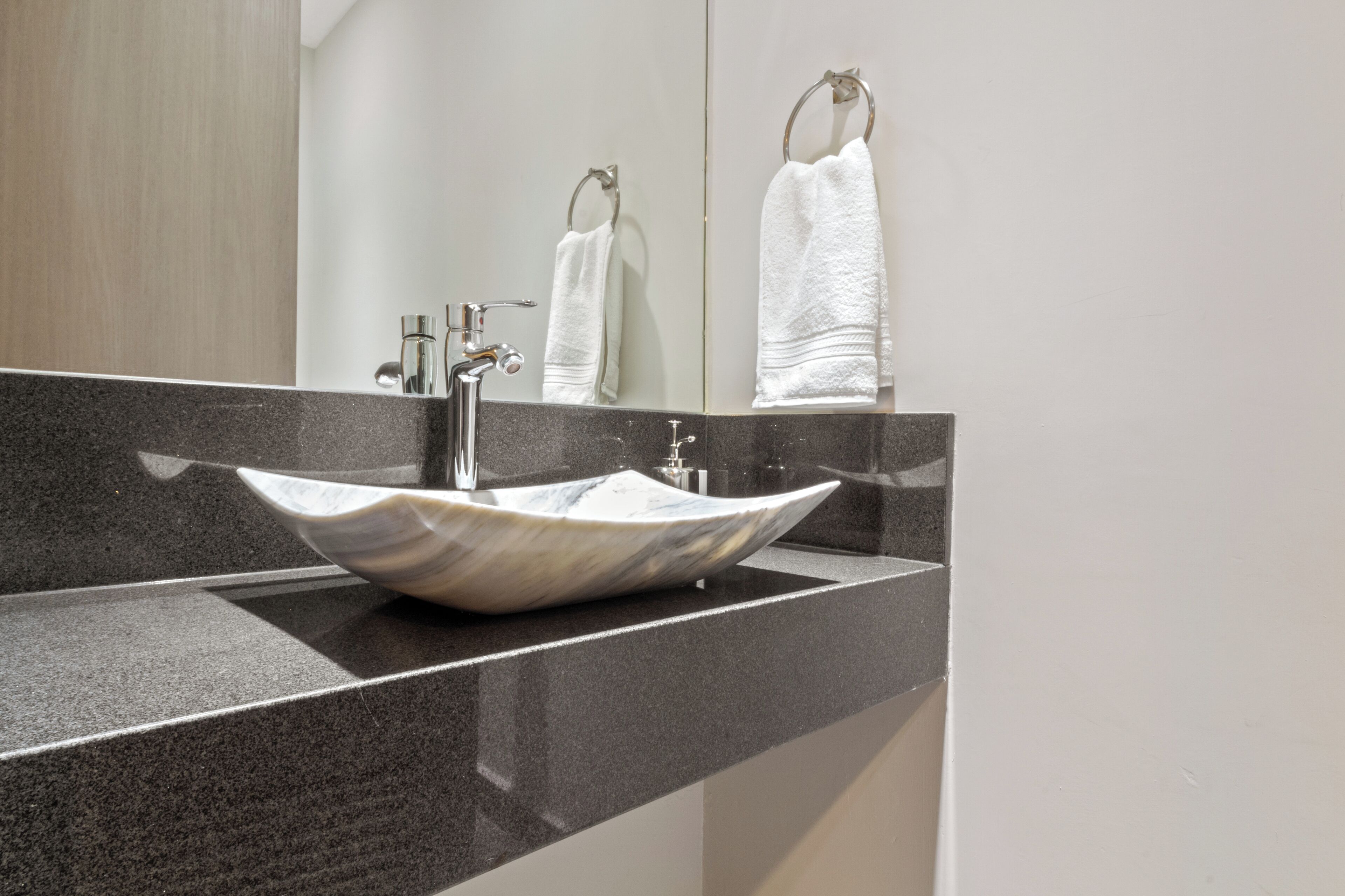 A modern bathroom with a large, white, marble vessel sink, a chrome faucet, and a mirror reflecting a towel rack and another towel.