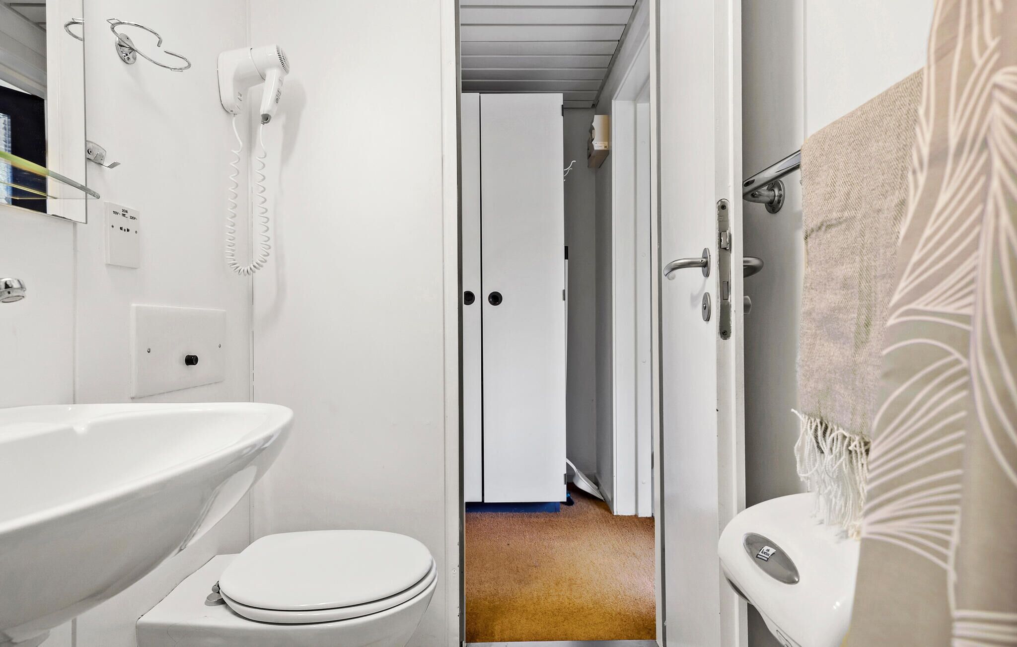 A bathroom with a white sink, toilet, and shower area. There is a hairdryer mounted on the wall and a towel rack with a towel.