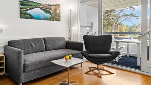 A modern living room with a grey sofa, a black armchair, a small white table with fruit, and a large window with a view of trees and a lake. - Amazing apartment in Nexø with WiFi (Nexø)