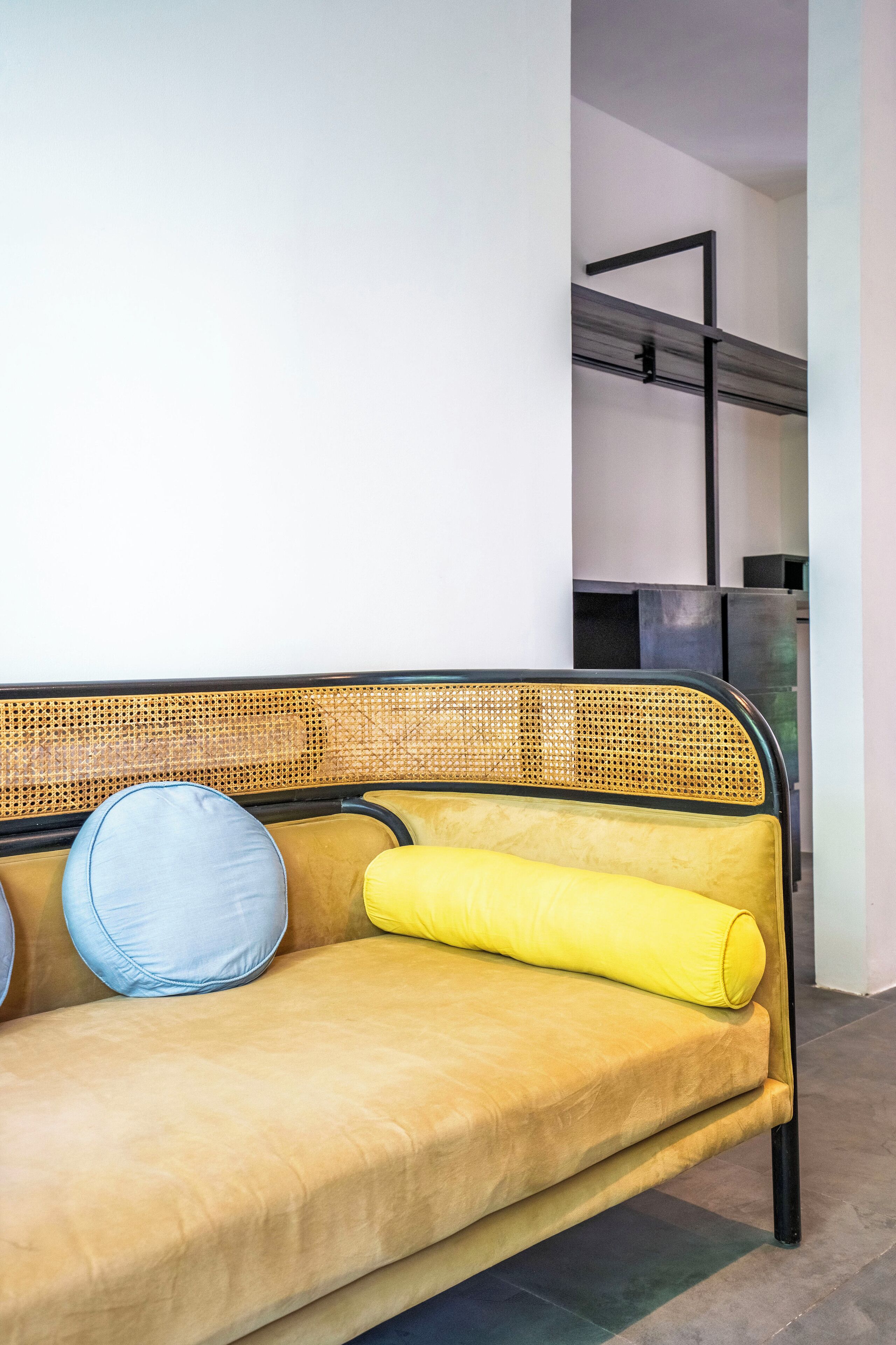 En gul sofa med en flettet ryg og to puder, en blå og en gul, i et rum med moderne design og synlig metalstruktur i baggrunden.