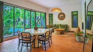 A spacious room with a large dining table, comfortable seating, a sofa, and a view of lush greenery through large windows. - Veronica Villa by Betterplace (Canggu)