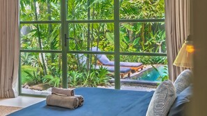 A bedroom with a large window, a bed with a blue bedspread, and a view of a pool and greenery outside. - Veronica Villa by Betterplace (Canggu)