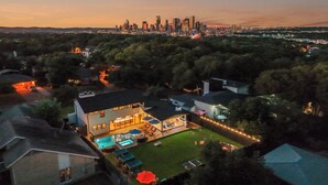 A residential area with a large house featuring a swimming pool, a patio, and a view of a city skyline at dusk. - Most EPIC Austin Experience! Sleeps 14 + heated Pool + Spa + Cabanas + Speakeasy (Austin)