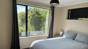 A bedroom with a large window, a bed with striped bedding, a bedside lamp, and a view of greenery outside. - Nature on your door step (Waitati)