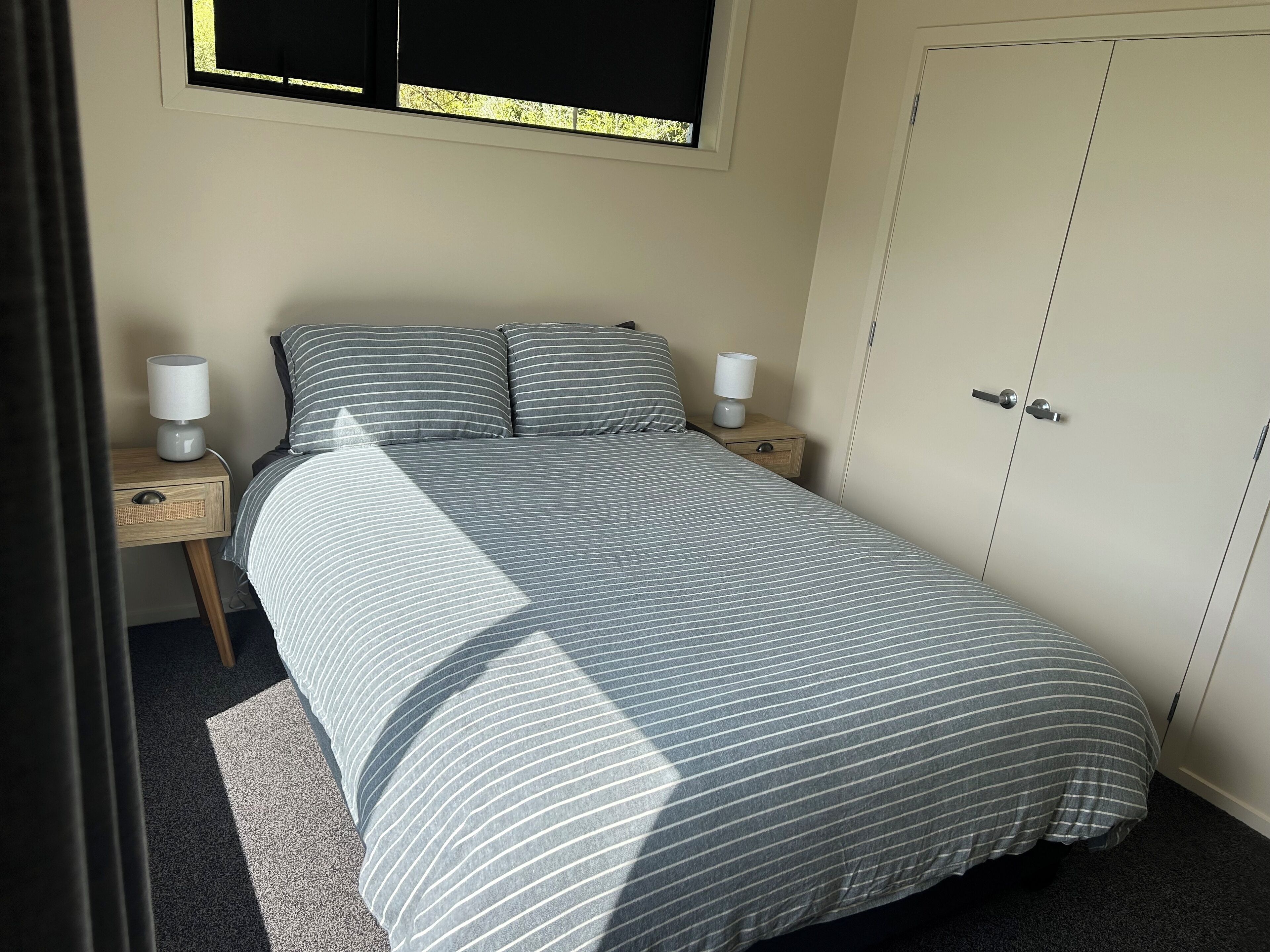 A neatly made bed with striped bedding, two bedside lamps, a small wooden nightstand, and a wardrobe with a door.