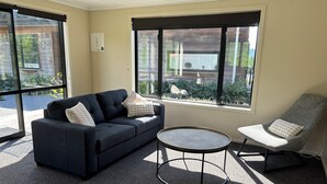 A modern room with a dark blue sofa, a grey armchair, and a small round table. Large windows provide a view of greenery outside. - Nature on your door step (Waitati)