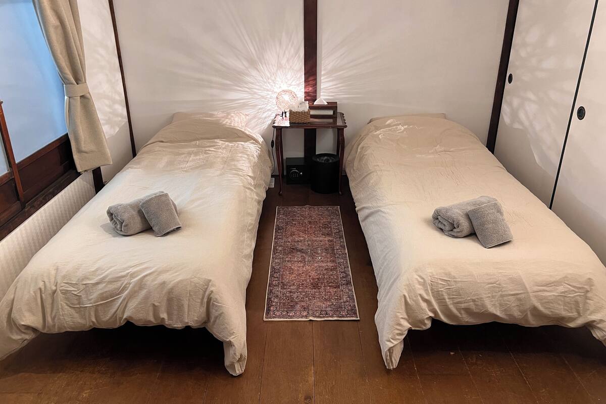 Una habitación con dos camas individuales, una mesita con lámpara, un tapete y una ventana con cortinas.