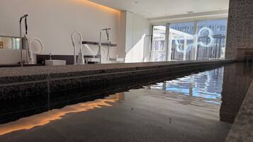 Un área de piscina interior moderna con una piscina larga y estrecha, una encimera de piedra y una barandilla de vidrio.