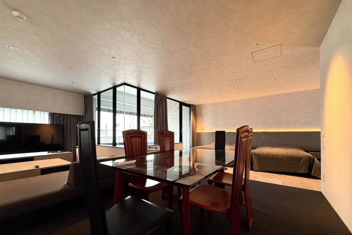 A modern hotel room with a glass dining table, wooden chairs, a flat-screen TV, a sofa, and a large window offering a city view.