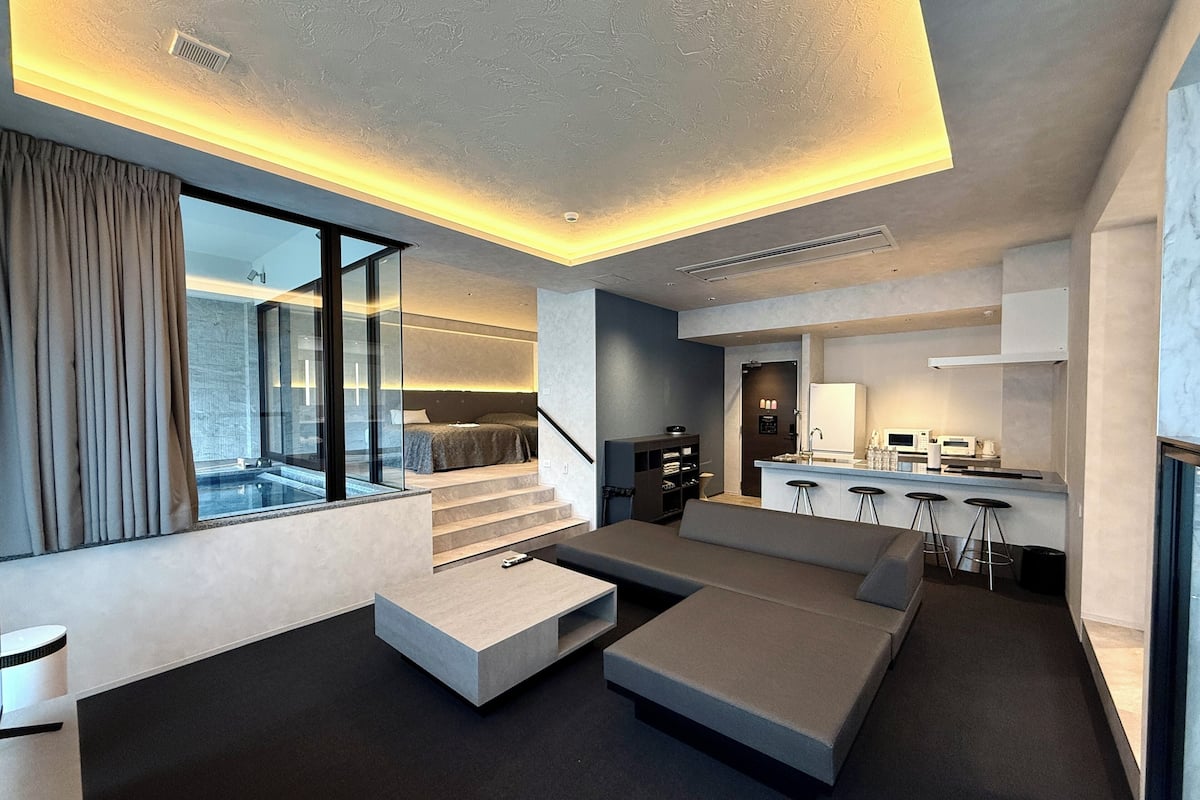 A modern living room with a large sectional sofa, a glass wall, and a kitchen area with bar stools.