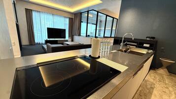 A modern kitchen with a built-in stove, a sink, and a countertop. There is a large window with curtains, a flat-screen TV, and a seating area with a sofa and a coffee table.