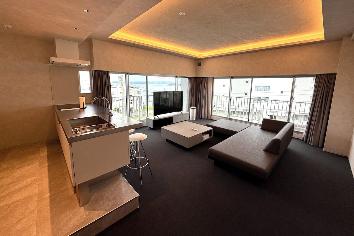 A modern living room with a kitchen area, large windows, and a balcony.