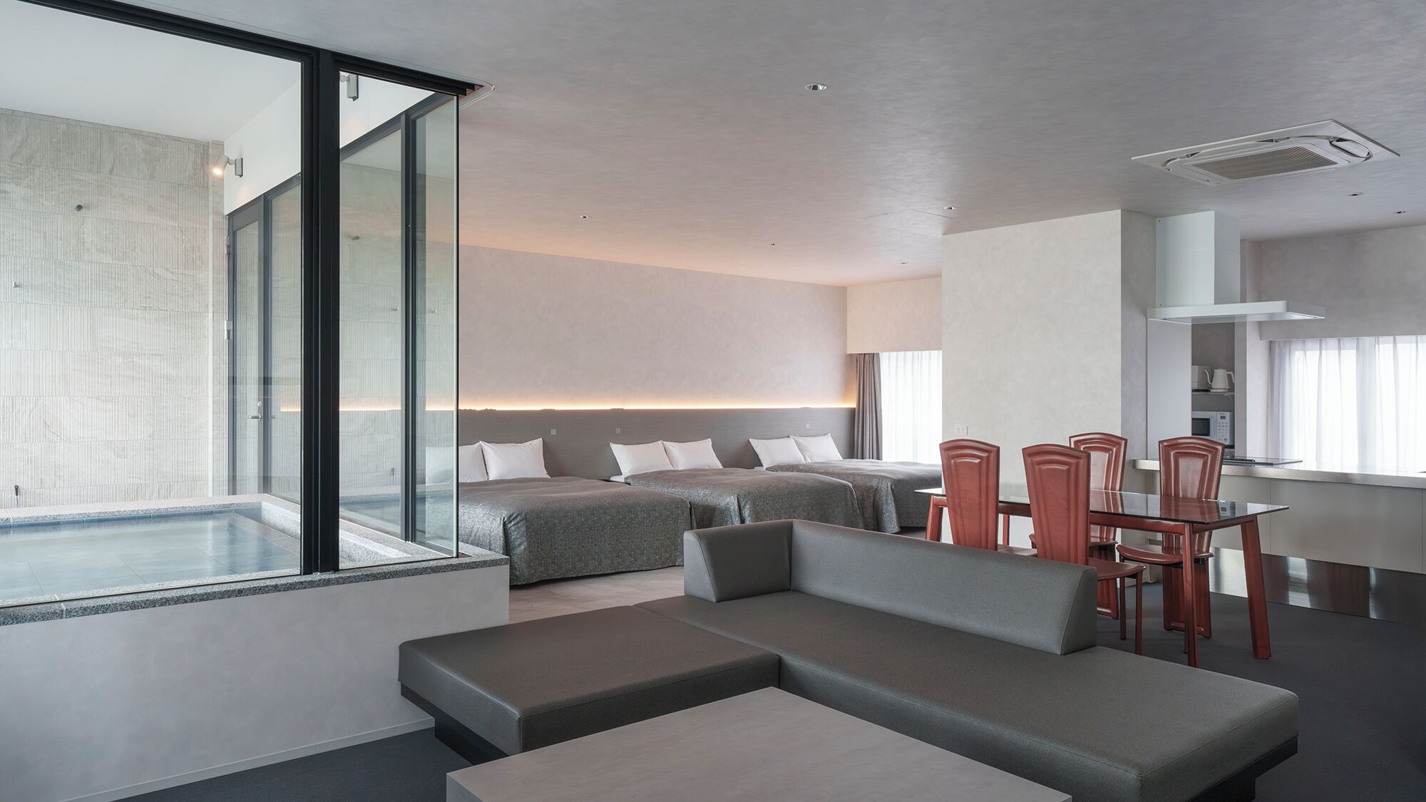 A modern hotel room with a glass wall, a large bed, a separate bed, a dining area, and a small kitchen.