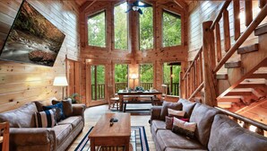 Un intérieur spacieux de chalet en bois, avec une grande table à manger, deux canapés et un escalier.