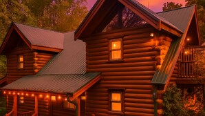 Une cabane en rondins dotée d’un porche et de nombreuses fenêtres, entourée de verdure.