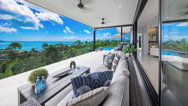 A modern outdoor lounge area with a view of the ocean, a pool, and a glass-enclosed living space.