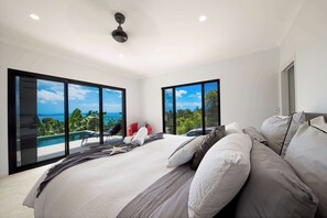 Un dormitorio con una cama grande, dos ventanas, un ventilador de techo y vista al océano.