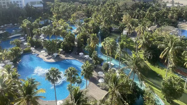 A resort with multiple swimming pools surrounded by palm trees and greenery, with a beach and mountains in the background.