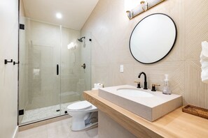Ein modernes Badezimmer mit einer in Glas eingefassten Dusche, einer weißen Toilette, einem großen Waschbecken mit Spiegel und einer hölzernen Ablagefläche.