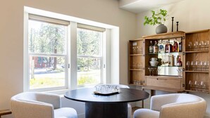 Un coin repas avec une table ronde et quatre chaises, un buffet en bois contenant de la verrerie, et une vue sur les arbres à travers de grandes fenêtres.