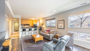 A modern living room with a grey sofa, a wooden coffee table, and a kitchen area with wooden cabinets and a bar stool.