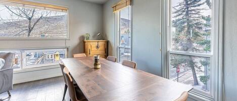 A dining area with a wooden table and chairs, a cabinet, and large windows with views of trees and buildings.
