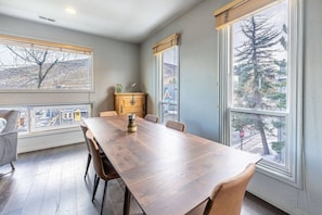 Un comedor con una mesa y sillas de madera, un armario y ventanas amplias con vistas a árboles y edificios.