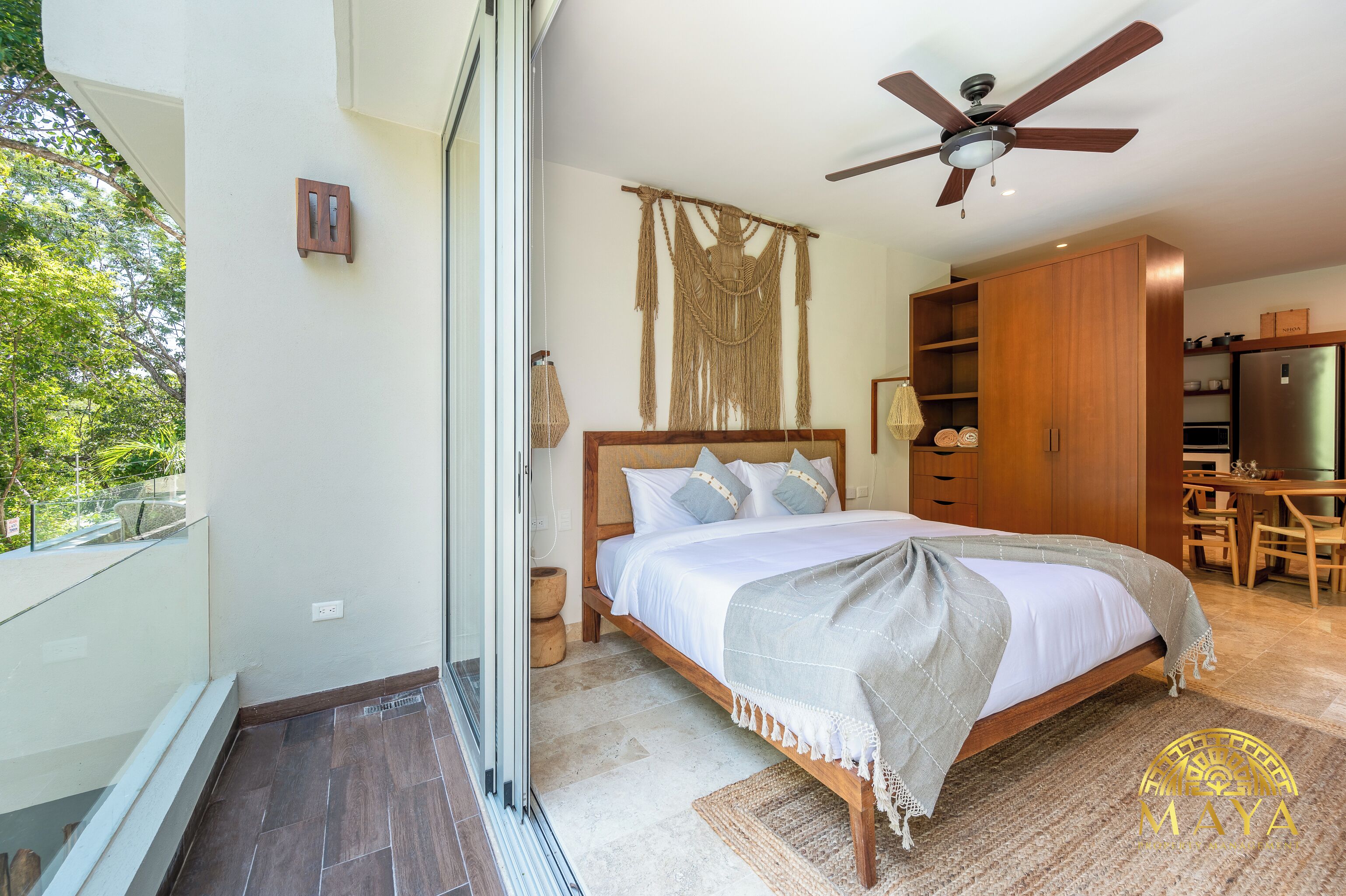 A bedroom with a bed, wooden wardrobe, ceiling fan, and a view of greenery outside.