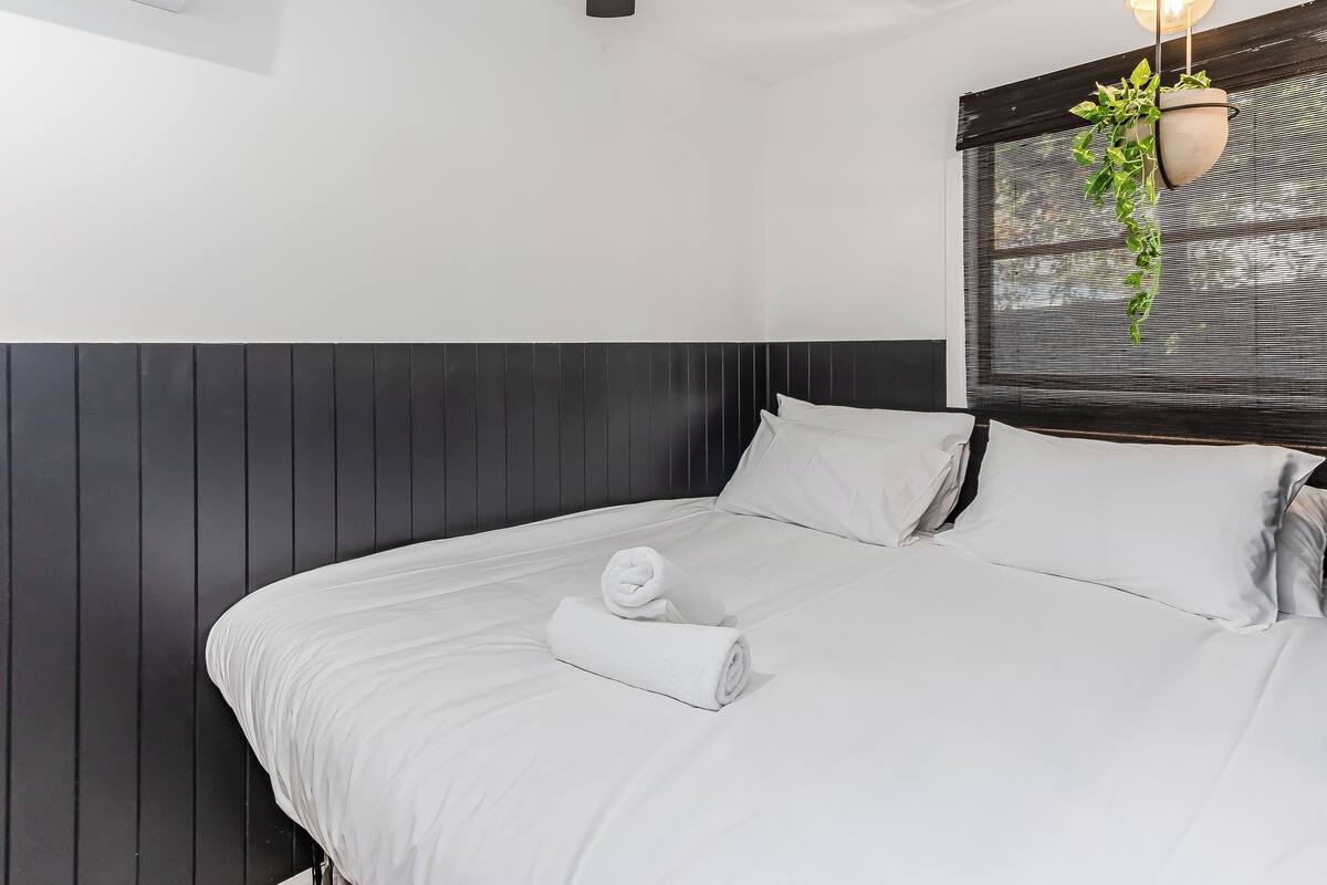 A neatly made bed with white linens, a rolled towel, and a potted plant hanging from the ceiling.