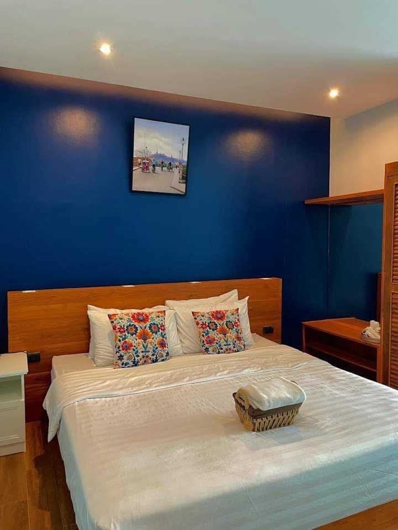 A bedroom with a wooden headboard, a bed with white bedding and colorful floral pillows, a bedside table with a lamp, and a wall-mounted picture.