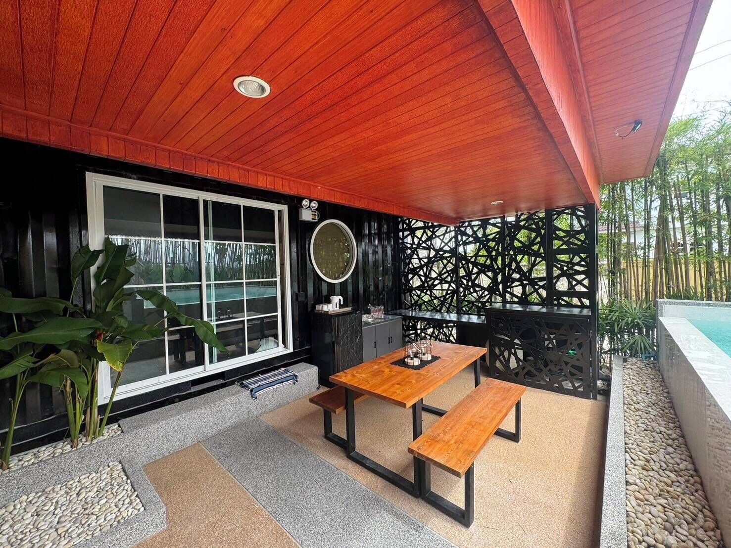 An outdoor patio with a wooden table and benches, a large window, and a decorative wall with a lattice design.