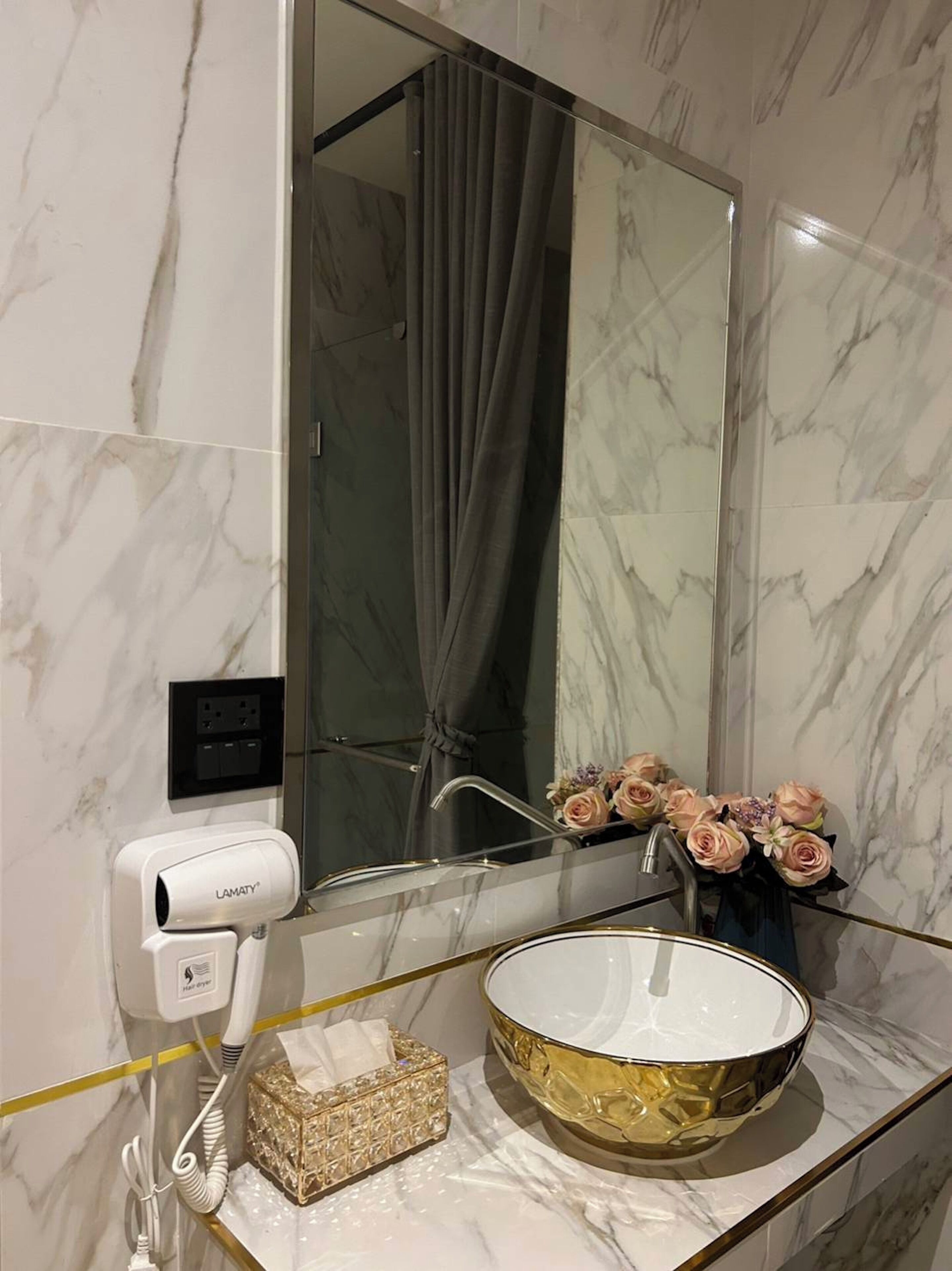 Un baño con lavamanos de mármol, un espejo grande, un secador de pelo y un florero con flores.