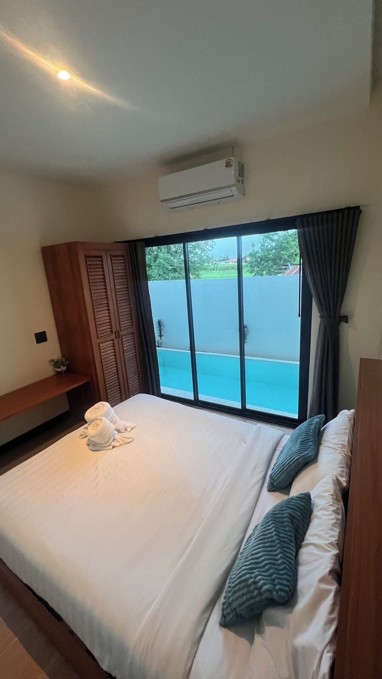A hotel room with a bed, a window with curtains, an air conditioning unit, and a view of a swimming pool.