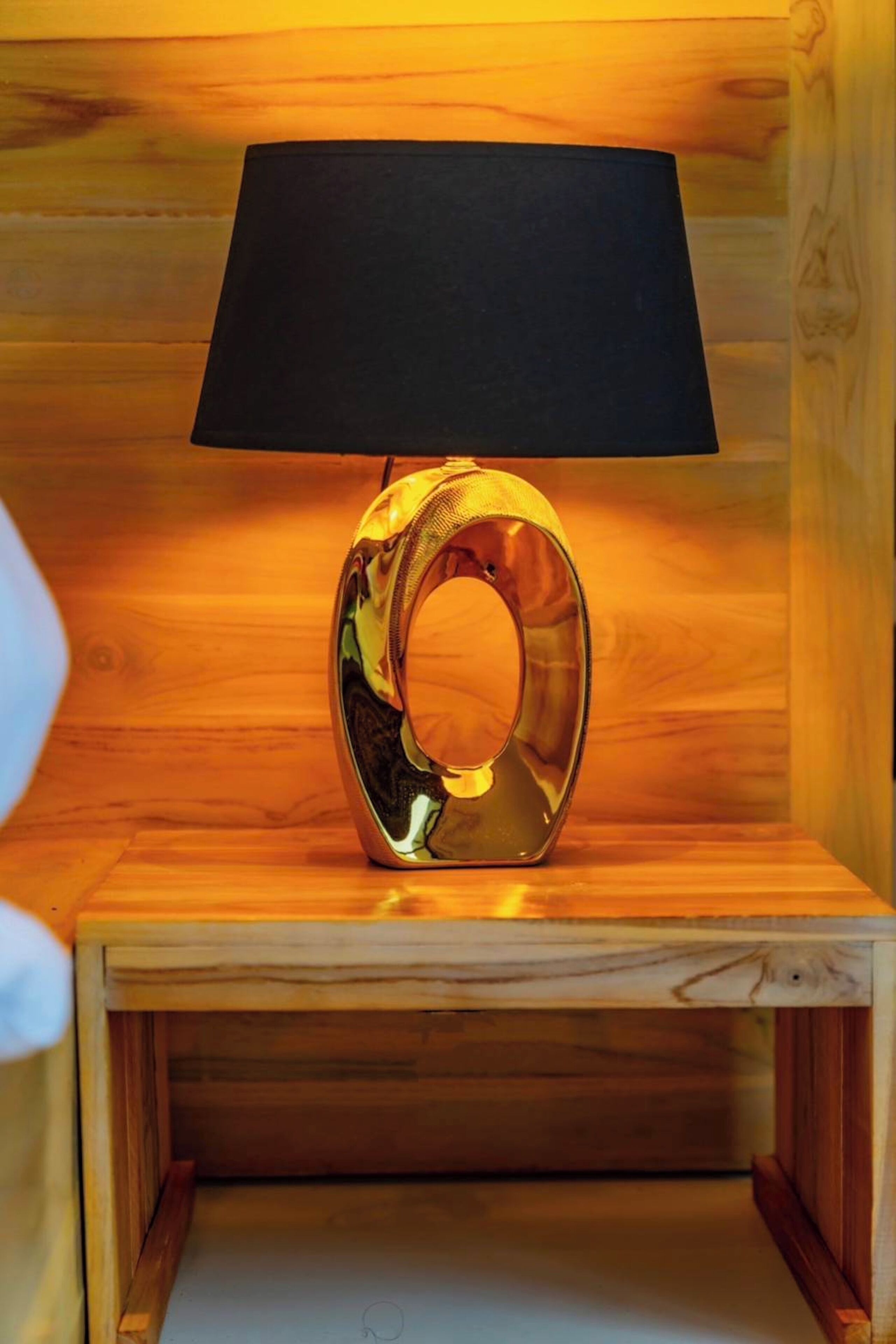 Eine moderne, metallische Lampe mit schwarzem Lampenschirm auf einem Nachttisch aus Holz.