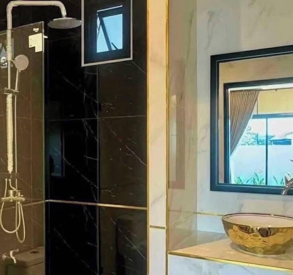 Un baño con pared de mármol negro, cabina de ducha de vidrio, espejo con marco dorado y una ventana con cortinas.